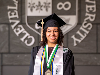 Leilah Flores posing in front of CSU seal wearing valedictorian stole and commencement regalia. 