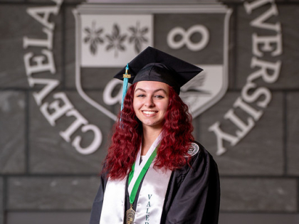 Vivian France posing in front of CSU seal wearing valedictorian stole and commencement regalia. 